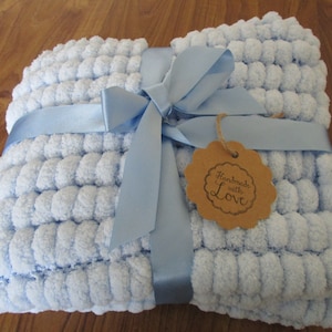 May include: A light blue baby blanket with a pom-pom texture, tied with a blue ribbon and a brown tag that says "Handmade with Love".