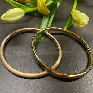 Peut inclure: Deux anneaux en métal couleur or, l'un légèrement plus grand que l'autre, sont posés sur une surface gris foncé. Les anneaux sont lisses et brillants, et ils sont positionnés de manière à se chevaucher légèrement.