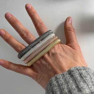 Puede incluir: Cuatro pulseras apiladas en diferentes colores metálicos. Las pulseras están hechas de pequeñas cuentas y se llevan en la muñeca.