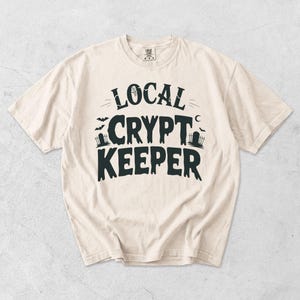 Peut inclure: Un t-shirt à manches courtes de couleur crème avec le texte noir LOCAL CRYPT KEEPER. Le motif comprend des éléments graphiques représentant une toile d'araignée, des chauves-souris, un croissant de lune et deux pierres tombales avec clôtures.