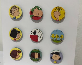Può includere: Una tavola quadrata bianca con nove magneti circolari raffiguranti i personaggi dei Peanuts disposti in una griglia tre per tre. I magneti mostrano vari personaggi, tra cui Snoopy e Woodstock, in illustrazioni colorate.
