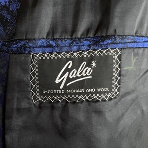 May include: A black garment interior featuring a rectangular black label with white text that reads Gala and IMPORTED MOHAIR AND WOOL. The label is stitched with white thread onto a black lining, adjacent to a blue and black patterned fabric.