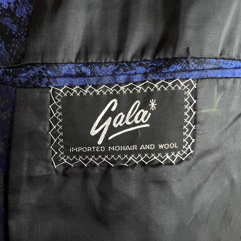 May include: A black garment interior featuring a rectangular black label with white text that reads Gala and IMPORTED MOHAIR AND WOOL. The label is stitched with white thread onto a black lining, adjacent to a blue and black patterned fabric.