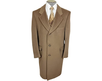 May include: A tan wool-blend overcoat featuring a notched lapel, three-button front closure, and two side flap pockets. The coat is worn over a white collared shirt and a tan patterned necktie, creating a structured and layered appearance.