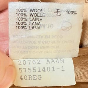 Op de afbeelding: Een wit stoffen waslabel met zwarte tekst die 100% wol aangeeft in meerdere talen, waaronder Engels, Duits, Frans, Italiaans en Spaans, samen met het Japanse kanji voor wol. Het bevat ook de tekst 20762 AA4M 57551401-1 40REG.