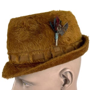 May include: A brown fedora hat with a textured, fur-like appearance. The hat features a decorative feather pin with red, blue, and gray feathers. The hat has a brown band around the crown and a brim.