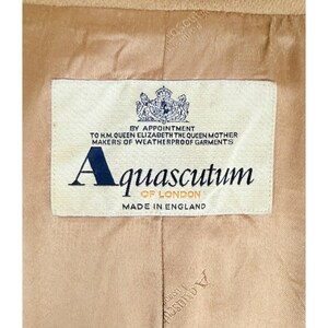 Op de afbeelding: De binnenkant van een beige kledingstuk met een rechthoekig wit stoffen label met de tekst BY APPOINTMENT TO H.M. QUEEN ELIZABETH THE QUEEN MOTHER MAKERS OF WEATHERPROOF GARMENTS, Aquascutum OF LONDON en MADE IN ENGLAND met een wapenschild.