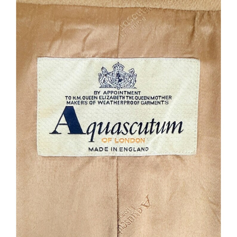 Op de afbeelding: De binnenkant van een beige kledingstuk met een rechthoekig wit stoffen label met de tekst BY APPOINTMENT TO H.M. QUEEN ELIZABETH THE QUEEN MOTHER MAKERS OF WEATHERPROOF GARMENTS, Aquascutum OF LONDON en MADE IN ENGLAND met een wapenschild.