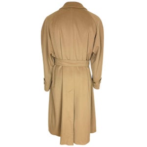 Op de afbeelding: Een lange, beige jas met een brede kraag, lange mouwen met knoopmanchetten en een bijpassende strikceintuur in de taille. De jas heeft een gestructureerd achterpand met een centrale naad en een enkele split aan de onderkant.
