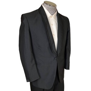 May include: A black dinner jacket featuring a shawl collar with black braided trim, a single fabric-covered button closure, and long sleeves with buttoned cuffs, worn over a cream-coloured button-down shirt with a grey chevron patterned collar.