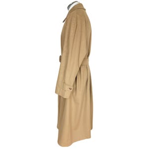 Op de afbeelding: Een lange, beige overjas met een klassieke kraag, lange mouwen met knoopmanchetten en een bijpassende stoffen riem in de taille. De jas wordt getoond in zijaanzicht en heeft een gladde, effen stof en een gestructureerd silhouet.