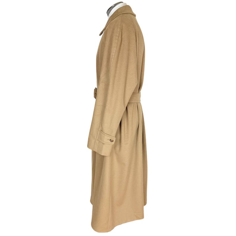 Op de afbeelding: Een lange, beige overjas met een klassieke kraag, lange mouwen met knoopmanchetten en een bijpassende stoffen riem in de taille. De jas wordt getoond in zijaanzicht en heeft een gladde, effen stof en een gestructureerd silhouet.