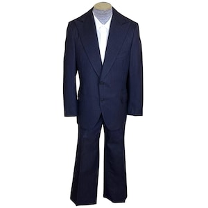 May include: A dark navy blue suit with thin vertical pinstripes, featuring a two-button blazer with wide lapels and matching flared trousers. The suit is worn over a crisp white collared shirt and a light grey patterned necktie.