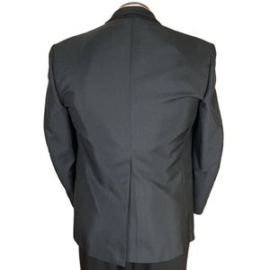 May include: A black blazer viewed from the back, featuring a structured collar with a subtle braided trim detail along the neckline. The garment has long sleeves, a central vertical seam, and a single vent at the bottom hem, showcasing a smooth fabric.