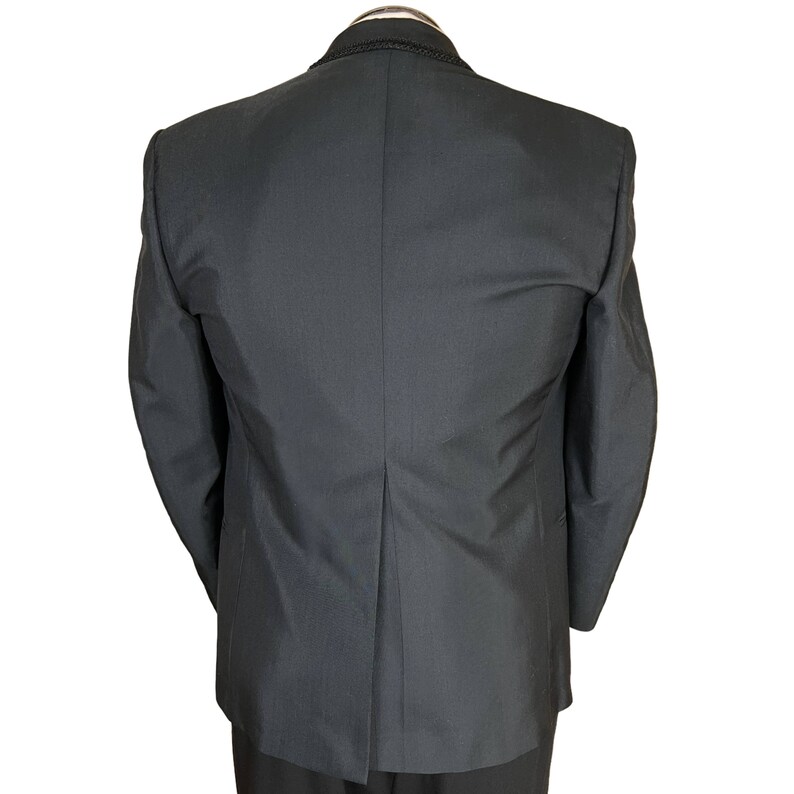 May include: A black blazer viewed from the back, featuring a structured collar with a subtle braided trim detail along the neckline. The garment has long sleeves, a central vertical seam, and a single vent at the bottom hem, showcasing a smooth fabric.