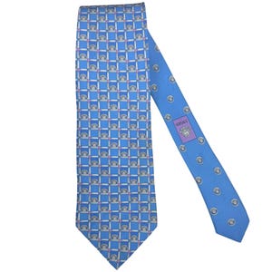 Puede incluir: Una corbata azul de seda con un patrón repetitivo de pequeños arcos y cuadrados. La corbata presenta un fondo azul claro con un diseño geométrico en blanco, rosa y gris. El logotipo de Versace es visible en la parte posterior de la corbata.