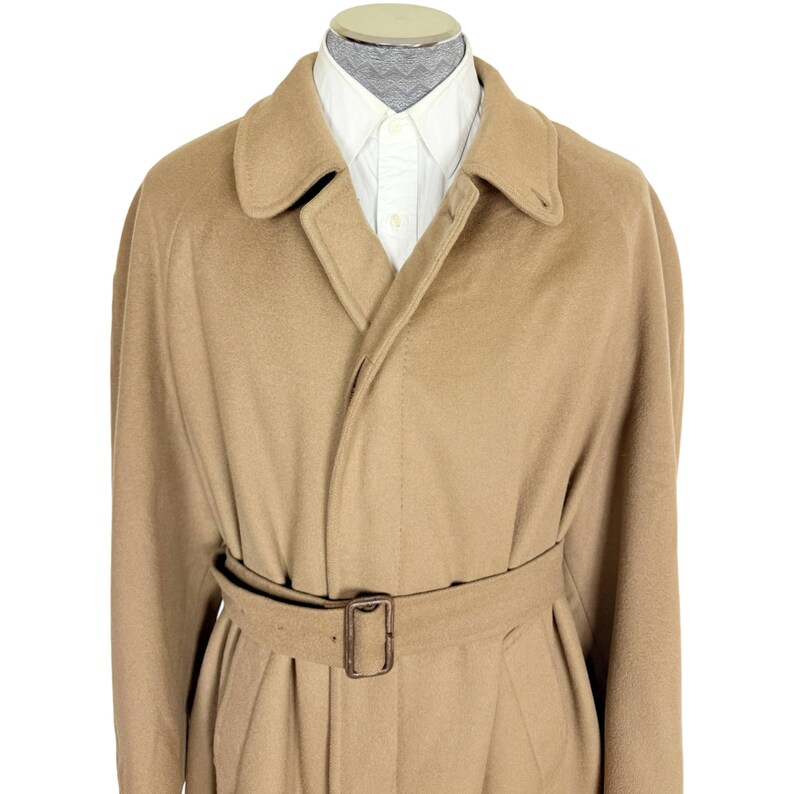 Op de afbeelding: Een beige overjas met een brede kraag, lange mouwen en een bijpassende stoffen riem met een rechthoekige gesp. De jas wordt gedragen over een wit overhemd met een zichtbare kraag, wat zorgt voor een gestructureerd en klassiek silhouet.