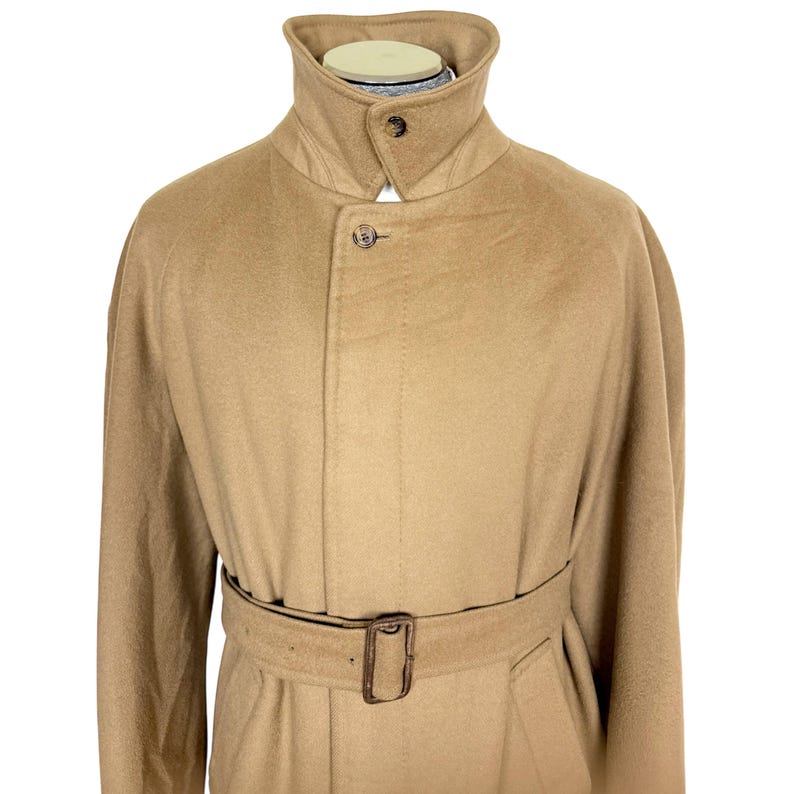 Op de afbeelding: Een beige jas van wolmix met een hoge opstaande kraag met &eacute;&eacute;n knoopsluiting, een verborgen sluiting aan de voorzijde met een zichtbare bovenste knoop en een bijpassende ceintuur met een rechthoekige gesp.