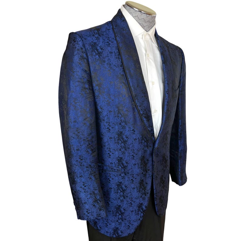 May include: A blazer featuring a vibrant blue and black mottled pattern with a shawl collar and a single button closure. The garment is worn over a crisp white button-down shirt with a visible collar, showcasing a structured fit and long sleeves.