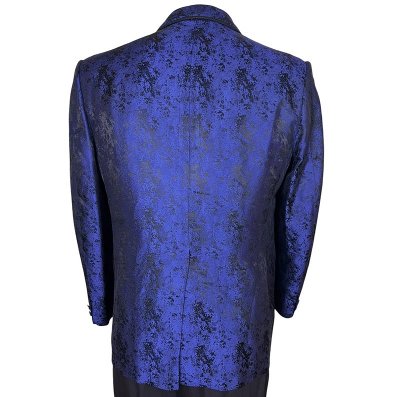 May include: A long-sleeved blazer in a vibrant royal blue hue, featuring a dark black mottled or abstract pattern throughout the fabric. The garment is shown from the back, displaying a structured collar, shoulder seams, and a central vertical seam.