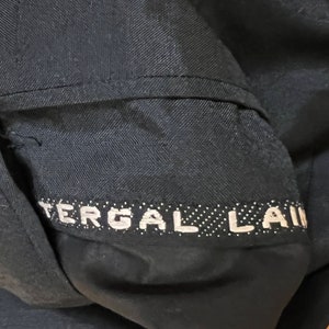 May include: A dark navy blue fabric garment featuring a horizontal band with the white text TERGAL LAINE. The material displays a visible herringbone weave pattern, and the text is set against a dark background with small white dots.