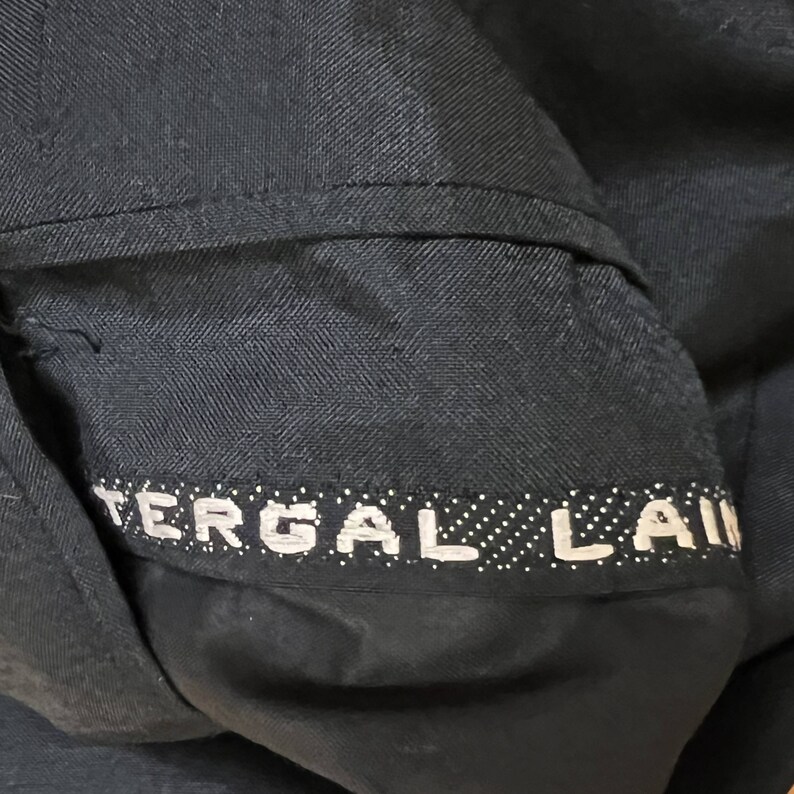 May include: A dark navy blue fabric garment featuring a horizontal band with the white text TERGAL LAINE. The material displays a visible herringbone weave pattern, and the text is set against a dark background with small white dots.