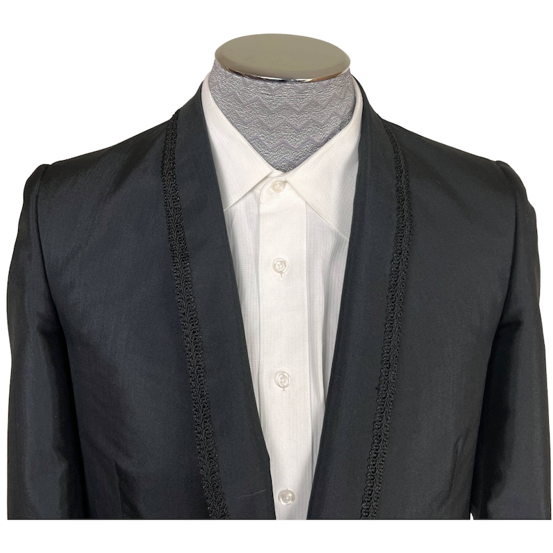 May include: A black blazer featuring a shawl collar with decorative black braided trim, worn over a crisp white button-down shirt. The blazer has a smooth, slightly lustrous finish, while the white shirt displays a subtle vertical texture.