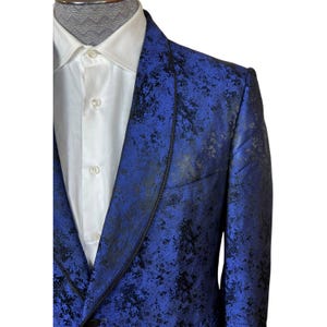 May include: A vibrant blue blazer featuring a dark, mottled black pattern throughout the fabric. The garment includes a shawl lapel with a thin black braided trim, a single-breasted front with buttons, and is worn over a crisp white collared shirt.