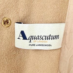 Op de afbeelding: Een beige kledingstuk van lamswol met een wit stoffen label met de tekst Aquascutum OF LONDON PURE LAMBSWOOL in zwarte en gouden letters. Een beige knoop met vier gaten is zichtbaar aan de linkerkant van het rechthoekige merklabel.