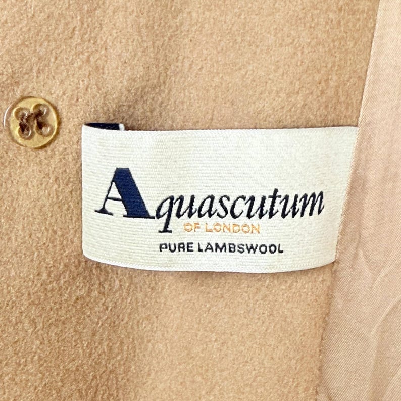Op de afbeelding: Een beige kledingstuk van lamswol met een wit stoffen label met de tekst Aquascutum OF LONDON PURE LAMBSWOOL in zwarte en gouden letters. Een beige knoop met vier gaten is zichtbaar aan de linkerkant van het rechthoekige merklabel.