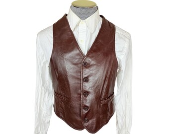 May include: A dark brown leather vest featuring a V-neckline, a five-button front closure, and two lower front welt pockets. The vest is layered over a crisp white long-sleeved button-down shirt with a classic collar and a light grey patterned necktie.