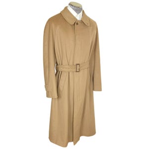 Op de afbeelding: Een beige lange jas met een puntige kraag, een knoopsluiting aan de voorzijde en een bijpassende stoffen riem met een rechthoekige gesp. De jas wordt getoond op een etalagepop die een wit overhemd onder de halslijn draagt.