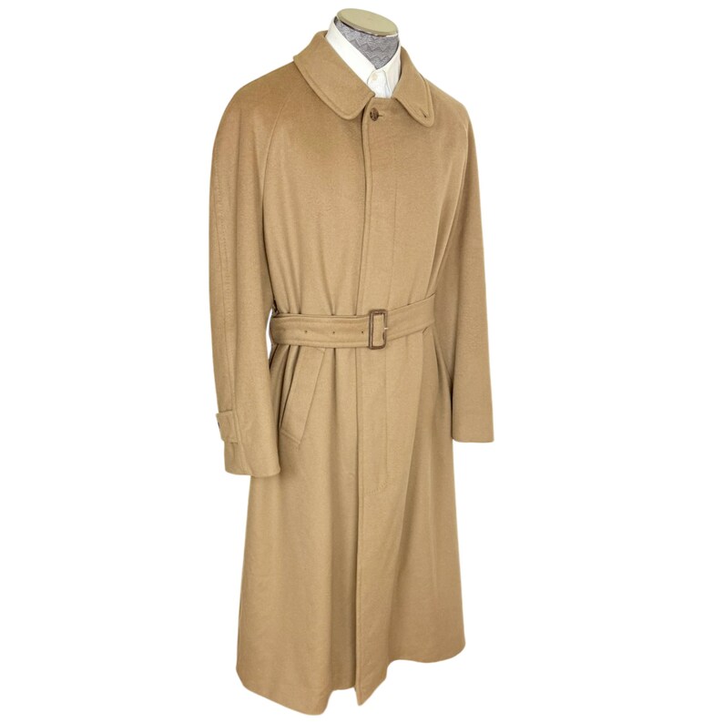 Op de afbeelding: Een beige lange jas met een puntige kraag, een knoopsluiting aan de voorzijde en een bijpassende stoffen riem met een rechthoekige gesp. De jas wordt getoond op een etalagepop die een wit overhemd onder de halslijn draagt.