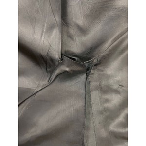 May include: A close-up view of black fabric lining featuring a visible seam and a small tear in the stitching. The material has a smooth, slightly reflective surface with natural folds and creases throughout the dark, solid-coloured textile.