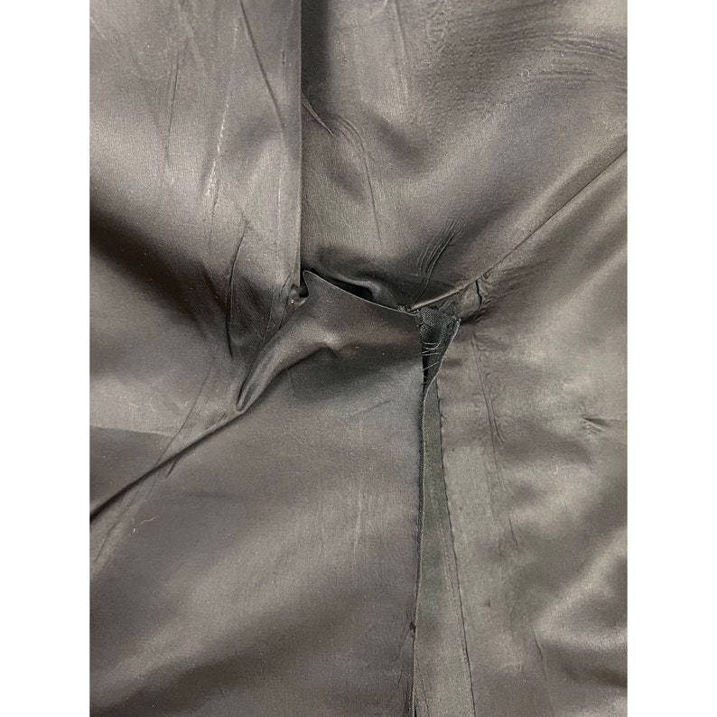 May include: A close-up view of black fabric lining featuring a visible seam and a small tear in the stitching. The material has a smooth, slightly reflective surface with natural folds and creases throughout the dark, solid-coloured textile.