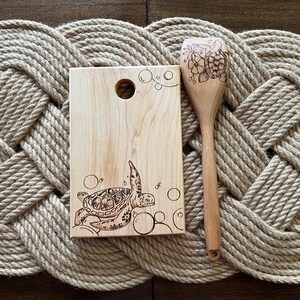 May include: A wooden cutting board and a wooden spoon with a sea turtle design burned into the wood. Both items are on a woven rope mat.