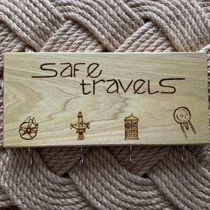 Pode incluir: Uma placa de madeira com o texto "safe travels" gravado na superfície. A placa tem quatro ganchos e apresenta quatro pequenas imagens de naves espaciais populares: o Millennium Falcon, um caça TIE, a TARDIS e a nave estelar Enterprise.