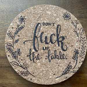 May include: A round cork coaster with a black floral design and the text "Don't Fuck Up The Fable."