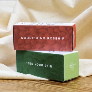 K&ouml;nnte beinhalten: Zwei kleine Boxen, die &uuml;bereinander gestapelt sind. Die obere Box ist rot mit wei&szlig;em Text, der "Nourishing Rosehip" lautet. Die untere Box ist gr&uuml;n mit wei&szlig;em Text, der "Feed Your Skin" lautet und einem kleinen Logo, das "GOODS FOR SKIN" lautet.