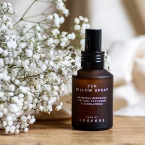 May include: A brown glass bottle with a black spray nozzle and a white label that reads "ZEN PILLOW SPRAY" with the ingredients listed below. The label also includes the text "MADE BY COOPERS".