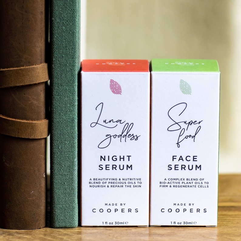 K&ouml;nnte beinhalten: Zwei kleine Flaschen mit Gesichts-Serum mit wei&szlig;en Etiketten und gr&uuml;nen und roten Akzenten. Die Flaschen sind mit "Luna goddess Night Serum" und "Super food Face Serum" beschriftet. Beide Flaschen werden von Coopers hergestellt und enthalten 1 fl oz (30 ml) Serum.