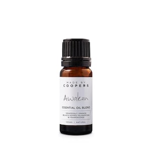Puede incluir: Una pequeña botella de vidrio ámbar con tapa negra. La etiqueta dice "MADE BY COOPERS Awaken ESSENTIAL OIL BLEND" con detalles de ingredientes: pomelo, naranja, pimienta negra, palmarosa e incienso. La parte inferior de la etiqueta dice "VEGAN | NATURAL".