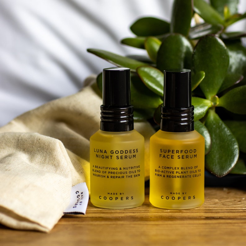 K&ouml;nnte beinhalten: Zwei bernsteinfarbene Glasflaschen mit schwarzen Kappen und Etiketten. Die Flaschen sind mit "Luna Goddess Night Serum" und "Superfood Face Serum" beschriftet. Die Etiketten enthalten auch den Text "Made by Coopers".