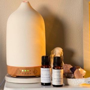 Puede incluir: Un difusor de aceites esenciales de color crema con base de madera y botones de control. Dos pequeñas botellas marrones de mezclas de aceites esenciales están frente al difusor. Una pequeña estatua blanca y cristales también están en la imagen.