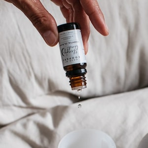 Puede incluir: Una botella de vidrio marrón con una etiqueta blanca que dice "MADE BY COOPER'S Sleepy (Moon) ESSENTIAL OIL BLEND VEGAN | LAVENDER | LEMONGRASS | YLANG YLANG" con una sola gota de aceite cayendo de la botella.
