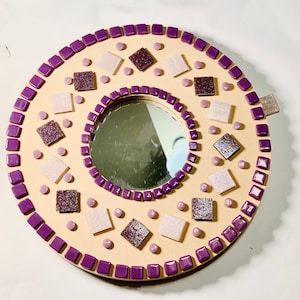 May include: A round mirror with a purple and beige mosaic design. The mirror is surrounded by a border of small purple squares and has a variety of beige and purple tiles in the center.