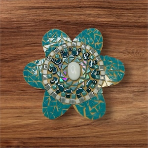 May include: A teal blue mosaic flower with a white center and a white pearl in the middle. The flower is made of small pieces of glass and is set against a wood background.