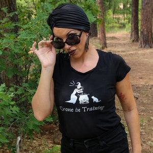 May include: A woman wearing a black turban, sunglasses, and a black t-shirt with the text "Crone in Training" and a graphic of a witch stirring a cauldron. She is wearing black denim shorts and is standing in a forest.