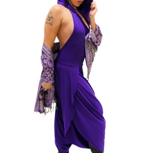 Op de afbeelding: Een vrouw draagt een paarse jumpsuit met capuchon en een paarse sjaal met zilveren patroon. Ze draagt zwarte laarzen en heeft een tatoeage op haar arm.