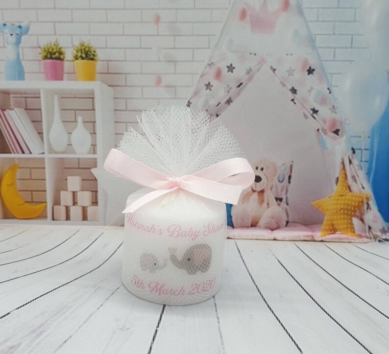 Puede incluir: Una vela blanca con un lazo y una cinta de tul rosa. La vela tiene un dise&ntilde;o rosa con dos elefantes y el texto "Hannah's Baby Shower 5th March 2021".